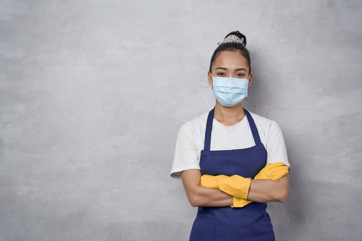 Confident cleaning lady wearing rubber gloves and medical protective face mask keeping arms crossed, looking at camera while standing against grey wall. Cleaning services during covid 19 pandemic