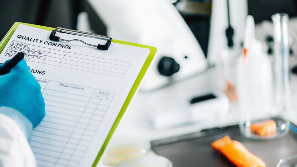 Gloved lab technician filling out a quality control inspection checklist in a research laboratory, illustrating documentation and contamination control as part of a structured research lab cleaning schedule.